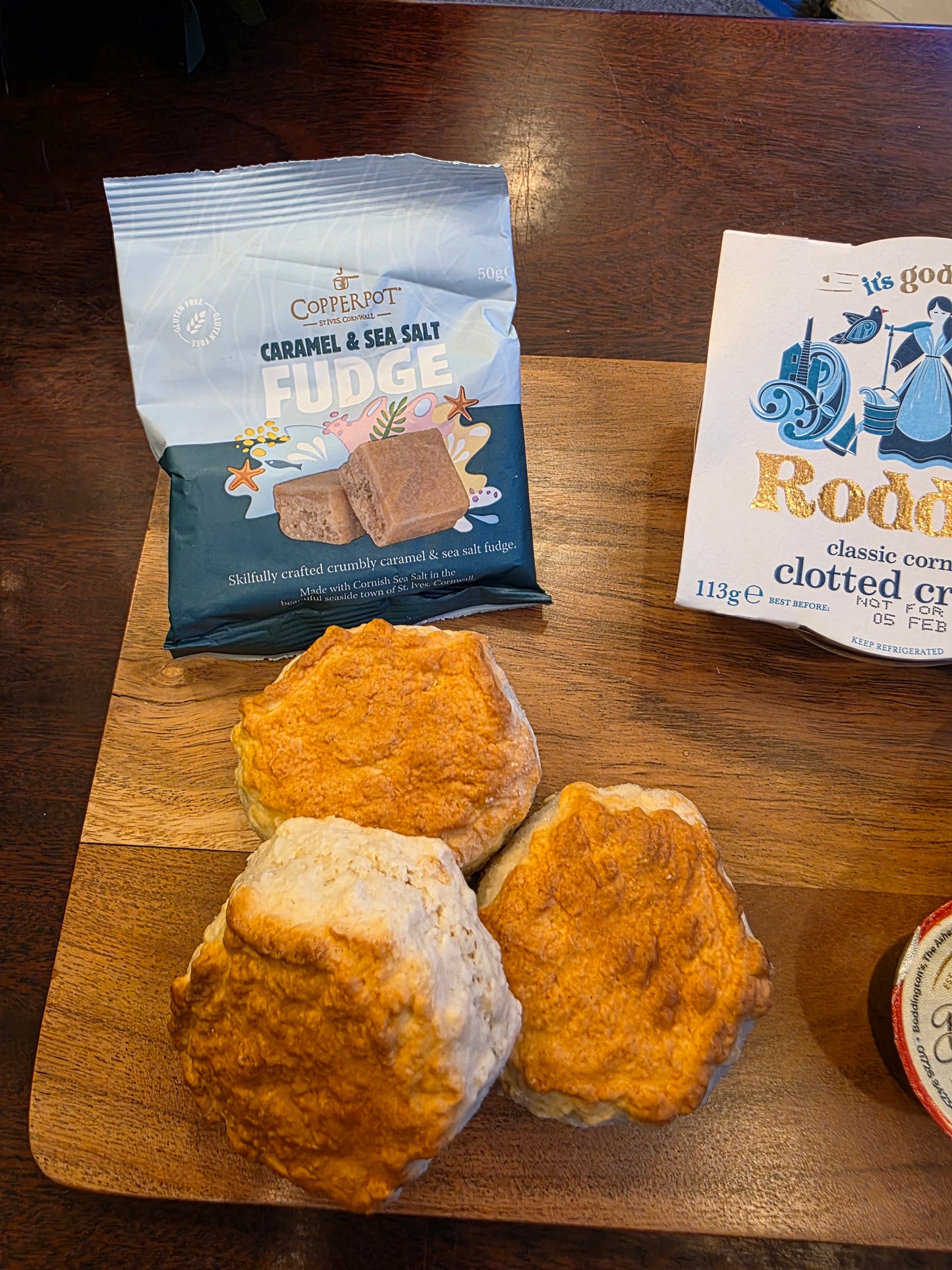 Three biscuits on a wooden board with a packet of fudge and a box of clotted cream in the background.