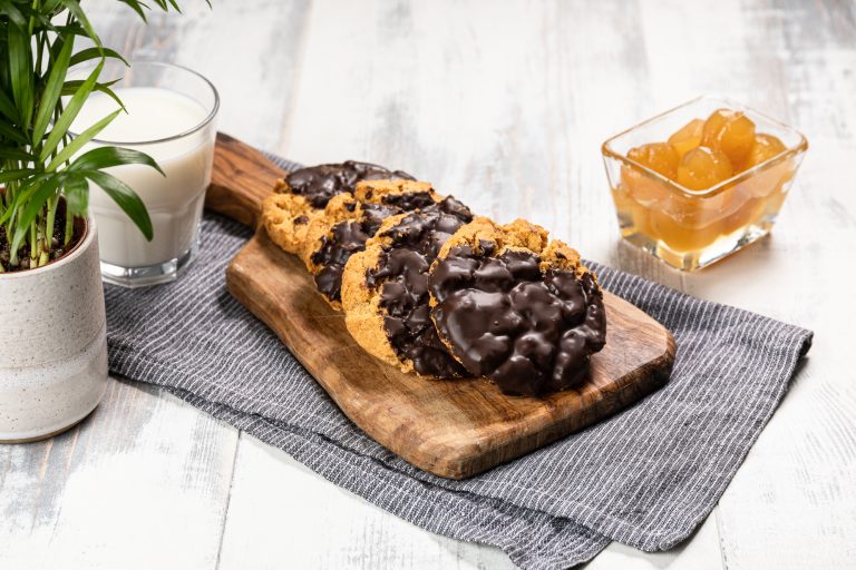 Chocolate chip cookies on a wooden board with a glass of milk and a small bowl of fruit in the background.