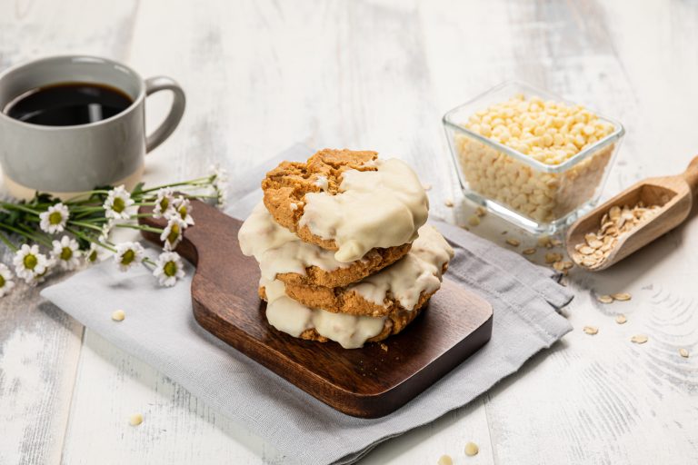 Two cookies with cream on a wooden board next to a cup of coffee and a bowl of white chocolate.