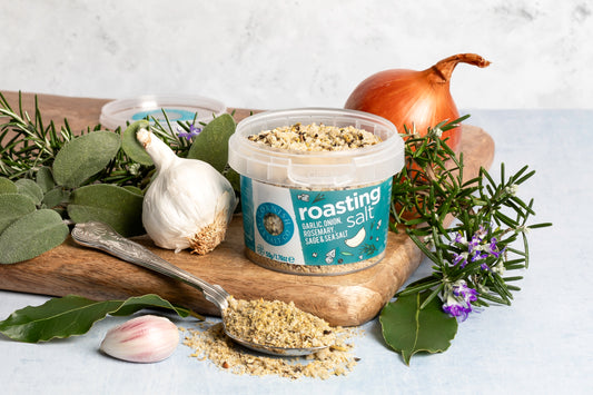 Container of 'Roasting Salt' with herbs, garlic, and an onion on a wooden board.