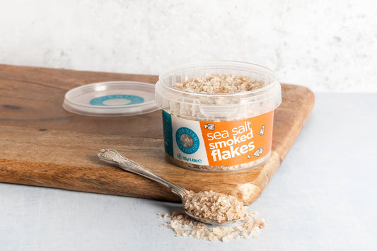 Jar of sea salt smoked flakes on a wooden board with a spoonful of flakes.