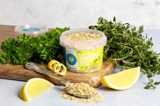 Container of 'freshly zesty' lemon salt with lemons, herbs, and a spoon on a wooden board.
