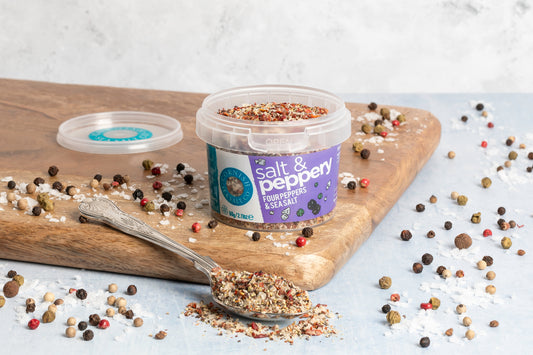 Container of salt and pepper mix on a wooden board with a spoonful of mix and scattered peppercorns.