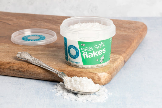 Container of sea salt flakes on a wooden board with a spoonful of salt next to it.
