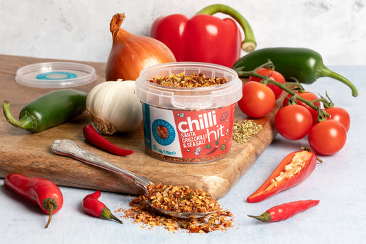 Container of Chilli Hit spice mix on a wooden board with chili peppers, tomatoes, and garlic.